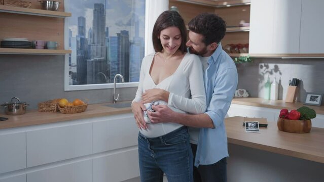 Caucasian young family of bearded husband with his pregnant wife embracing and holding little baby shoes. Aspiring parents. Affectionate couple. Kids concept.