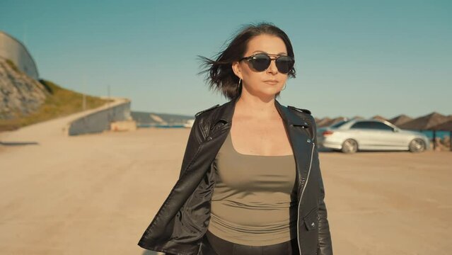 Portrait of independent woman in leather clothes holding a moto helmet and walking. Jib shot. Slow motion. The concept of girl power and moto travel.