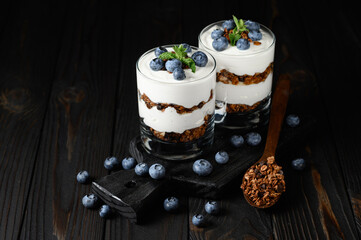Homemade layered dessert with fresh blueberries, cream cheese or yogurt, granola in glass on wood background.