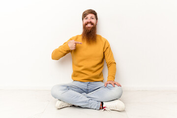 Young caucasian man sitting on the floor isolated on white background person pointing by hand to a shirt copy space, proud and confident