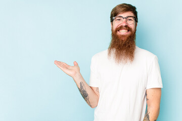 Young caucasian red-haired man isolated on blue background showing a copy space on a palm and holding another hand on waist.