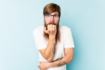 Young caucasian red-haired man isolated on blue background biting fingernails, nervous and very anxious.
