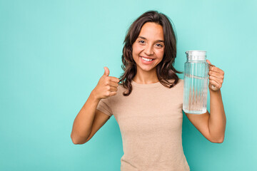 Young hispanic woman holding a water of jar isolated on blue background smiling and raising thumb up