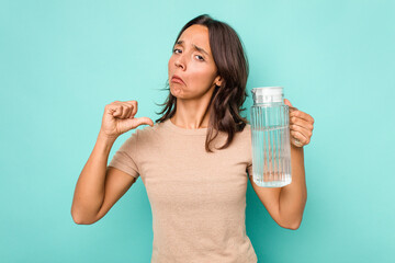 Young hispanic woman holding a water of jar isolated on blue background feels proud and self...