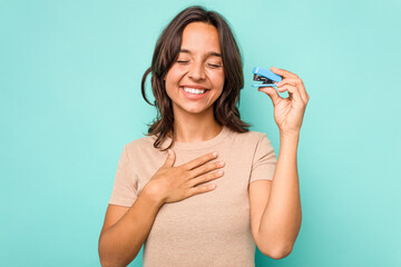 Young hispanic woman holding stapler isolated on blue background laughs out loudly keeping hand on...