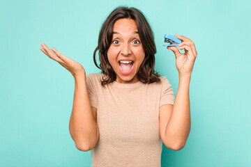 Young hispanic woman holding stapler isolated on blue background receiving a pleasant surprise,...