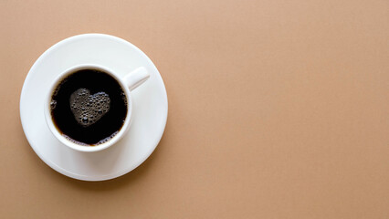 Top view of a cup of coffee on a saucer with a foam heart on a plain brown background with place...