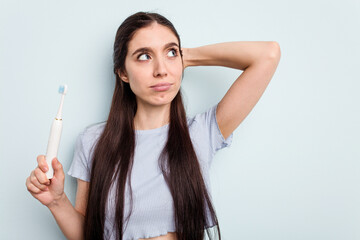 Young caucasian woman holding a electric toothbrush isolated on blue background touching back of head, thinking and making a choice.