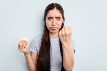 Young caucasian woman holding teeth whitener isolated on blue background showing fist to camera, aggressive facial expression.