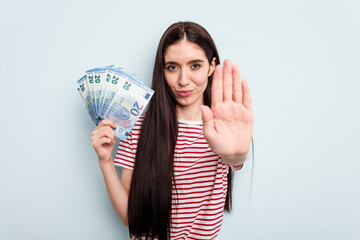 Young caucasian woman holding banknotes isolated on blue background standing with outstretched hand showing stop sign, preventing you.