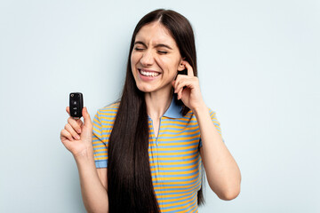 Young caucasian woman holding car keys isolated on blue background covering ears with hands.