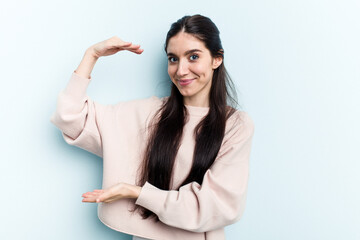 Young caucasian woman isolated on blue background holding something little with forefingers, smiling and confident.