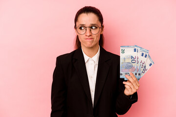 Young business caucasian woman holding bank notes isolated on pink background confused, feels doubtful and unsure.