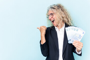 Middle age business caucasian woman holding bank notes isolated on white background points with thumb finger away, laughing and carefree.