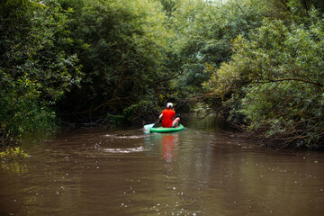 A narrow river among thickets of trees and bushes.. Girl surfer in red t-shirt sitting on the sup...