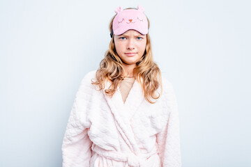 Caucasian teen girl wearing a pajama isolated on blue background shrugs shoulders and open eyes...