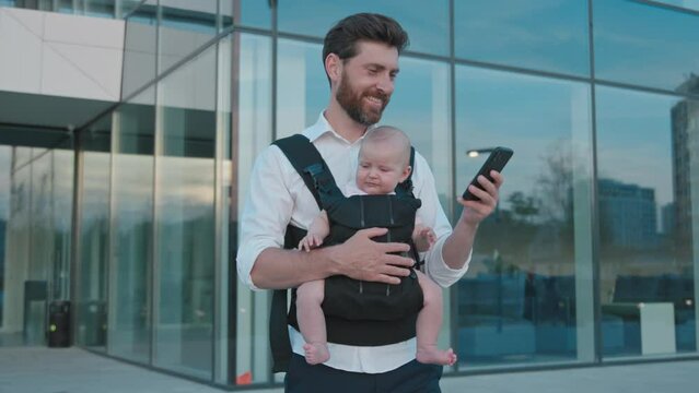 Young handsome father with a little newborn baby in a slings carrier walking and use phone typing smiling on street. Ergo backpack. Parents. Gadget. Smartphone. Cell. Slow motion
