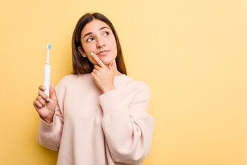Young caucasian woman holding a electric toothbrush isolated on yellow background looking sideways...