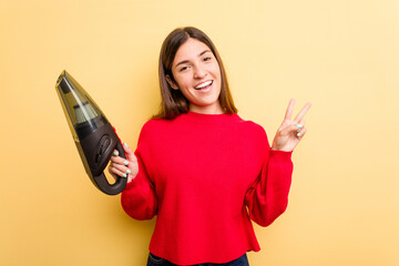 Young caucasian woman holding a hand vacuum cleaner isolated on yellow background joyful and carefree showing a peace symbol with fingers.