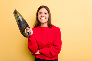 Young caucasian woman holding a hand vacuum cleaner isolated on yellow background laughing and having fun.