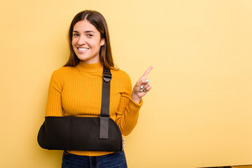 Young caucasian woman with broke arm isolated on yellow background smiling and pointing aside,...