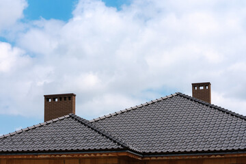 Dark metal tile shingles roof with chimney against blue sky with white clouds.