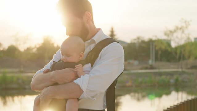 Portrait of attractive young father holding a child kiss and hug on street at sunlight funny newborn baby in a slings carrier. Ergo backpack. Parents. Slow motion