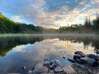 Paysage calme d'une forêt et d'une rivière à l'aube. Reflet d'une montagne sur un lac au matin. Lever de soleil avec brume sur l'eau. Rayons de soleil au travers le brouillard. Roches sur la rive.