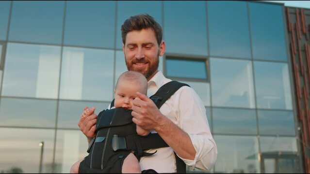 Cute portrait of funny newborn baby in a slings carrier. Young father holding a child kiss and hug on street. Ergo backpack. Parents. Slow motion