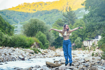 a beautiful tourist girl enjoys life near a mountain river on the rocks, in the background she has mountains