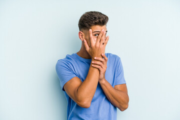 Young caucasian man isolated on blue background blink through fingers frightened and nervous.