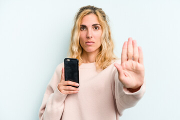 Fototapeta premium Young caucasian woman holding mobile phone isolated on blue background standing with outstretched hand showing stop sign, preventing you.