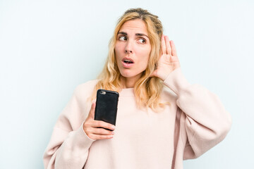 Naklejka premium Young caucasian woman holding mobile phone isolated on blue background trying to listening a gossip.