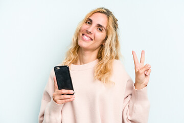 Fototapeta premium Young caucasian woman holding mobile phone isolated on blue background joyful and carefree showing a peace symbol with fingers.