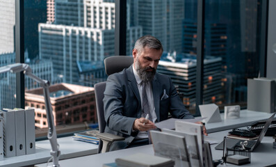 Fototapeta premium Portrait of 50s adult grey-haired Caucasian CEO businessman working at his desk in the office located in a skyscraper, business district buildings in the background