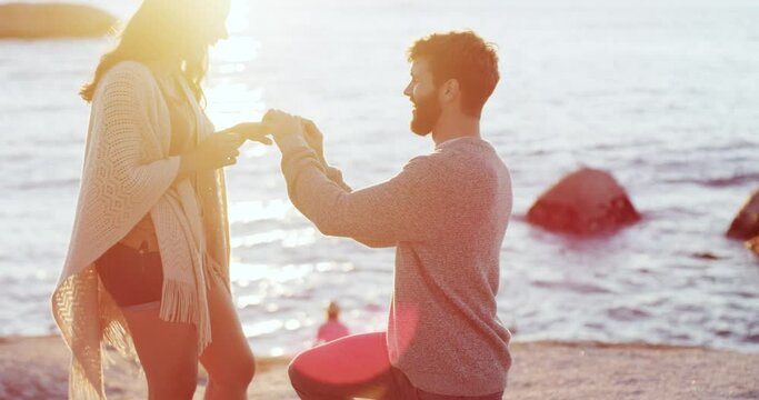 Romantic man proposing to woman on beach at sunset and hugging, embracing or holding his girlfriend after she says yes. Happy, smiling or excited couple getting engaged with a ring near sea or ocean
