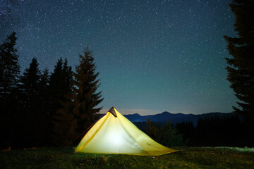 Bright illuminated tourist tent glowing on camping site in dark mountains under night sky with...