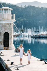 Fototapeta na wymiar Happy millenial mother and blonde teenager daughter in white dresses walking by the harbor and marina with yachts in a touristic sea resort with yachts and mountains