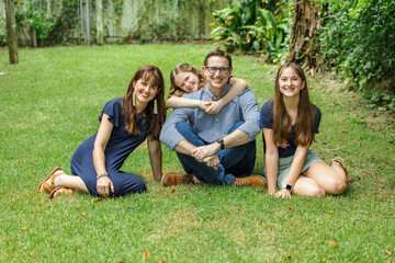 A family of a mother and father and two daughters sitting outside in the grass of their backyard witht the youngest daughter hugging dad from behind
