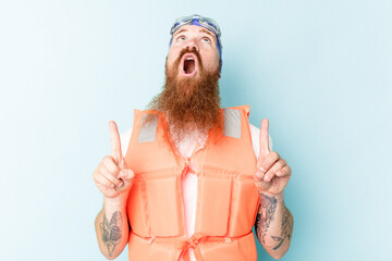 Young caucasian man wearing life jacket isolated on blue background pointing upside with opened mouth.