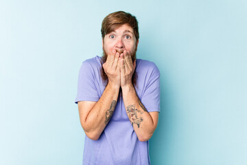Young caucasian red-haired man isolated on blue background shocked covering mouth with hands.