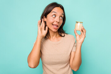 Young hispanic woman holding yogurt isolated on blue background trying to listening a gossip.