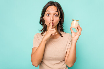 Young hispanic woman holding yogurt isolated on blue background keeping a secret or asking for silence.