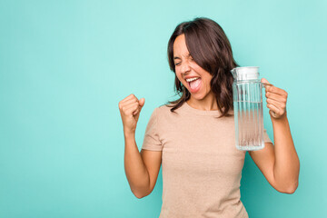 Young hispanic woman holding a water of jar isolated on blue background raising fist after a...