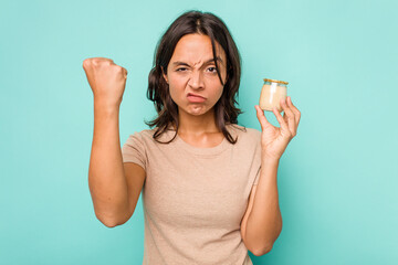 Young hispanic woman holding yogurt isolated on blue background showing fist to camera, aggressive...