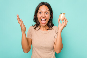 Young hispanic woman holding yogurt isolated on blue background receiving a pleasant surprise, excited and raising hands.