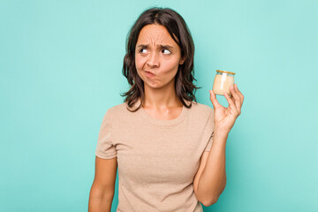 Young hispanic woman holding yogurt isolated on blue background confused, feels doubtful and unsure.