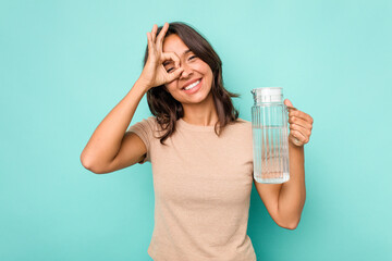 Young hispanic woman holding a water of jar isolated on blue background excited keeping ok gesture on eye.