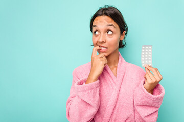 Young hispanic woman holding pills isolated on blue background relaxed thinking about something...