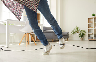 Man falls down as he stumbles over a power cable at home. Clumsy guy in sneakers trips over an electrical cord while walking on the floor in the living room. Cropped shot. Domestic accident concept - obrazy, fototapety, plakaty